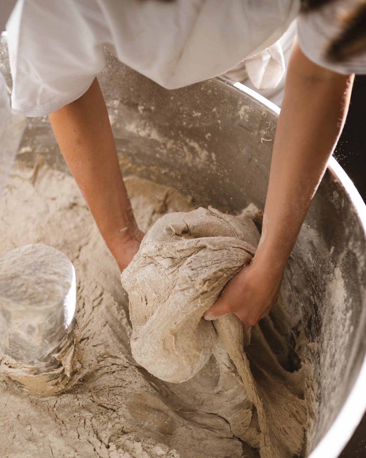 Eine Person knetet Teig in einer großen silbernen Schüssel. Die Hände sind mit Mehl bedeckt und der Teig hat eine grobe, unregelmäßige Form.