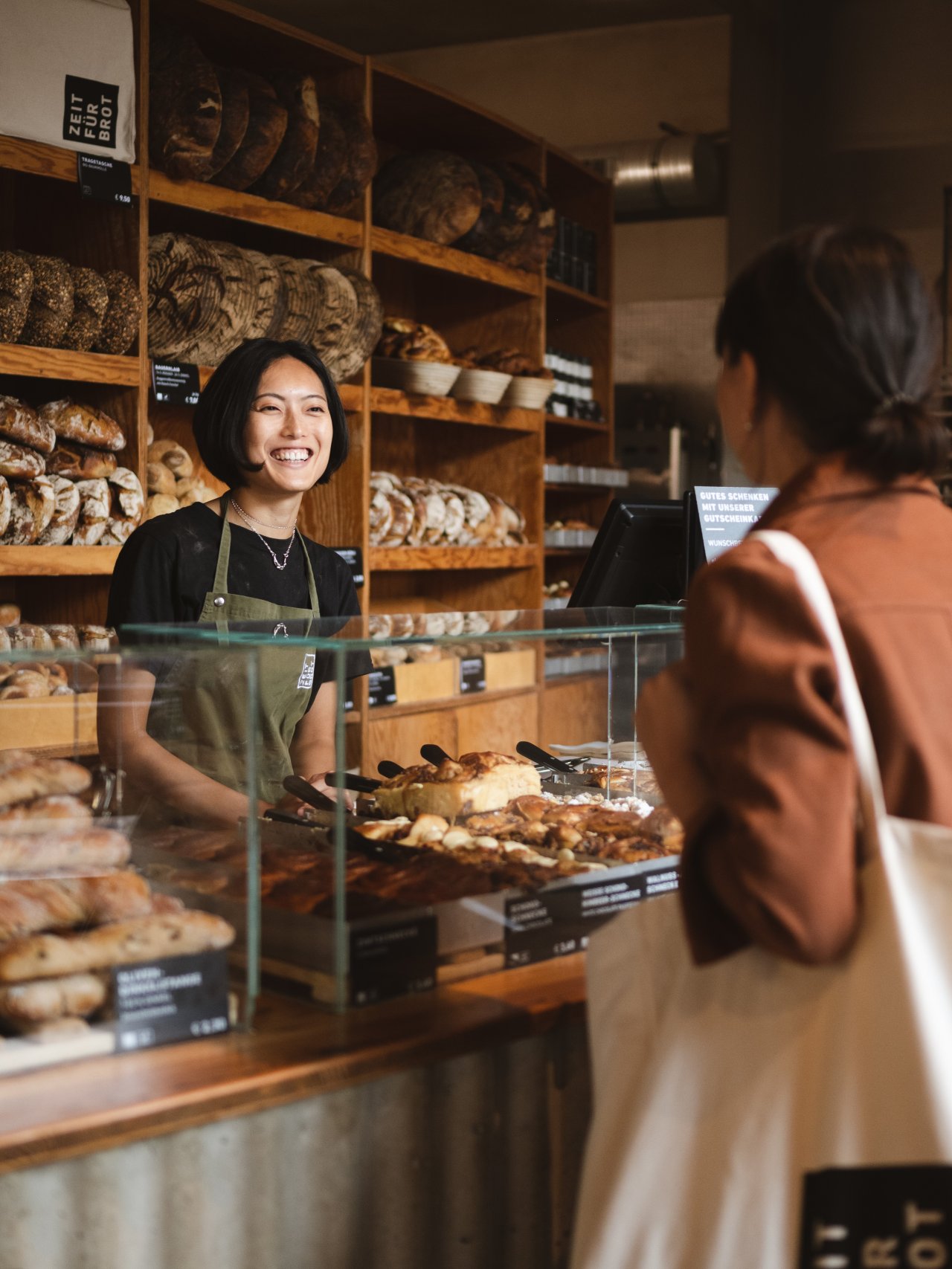 Eine lachende Person steht hinter einer Glastheke, umgeben von verschiedenen Broten und Gebäck. Sie trägt eine Grüne Verkaufsschürze. Eine Kundin mit einer hellen Tasche und roter Jacke schaut zur Verkäuferin.