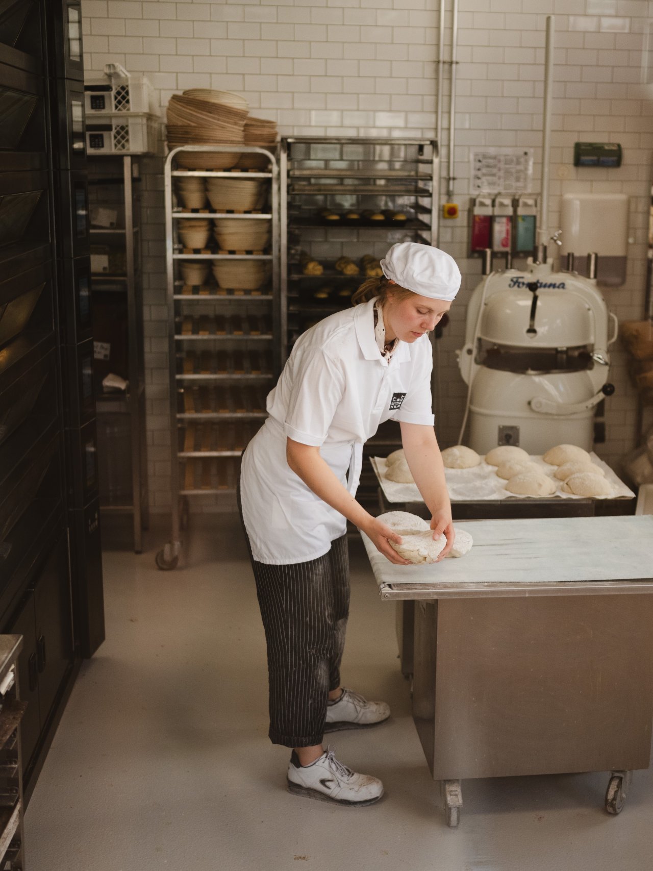 Eine Person in einem weißen T-Shirt und einer Schürze legt Brot auf ein Backblech. Im Hintergrund stehen Regale mit Schalen und ein Knetgerät.
